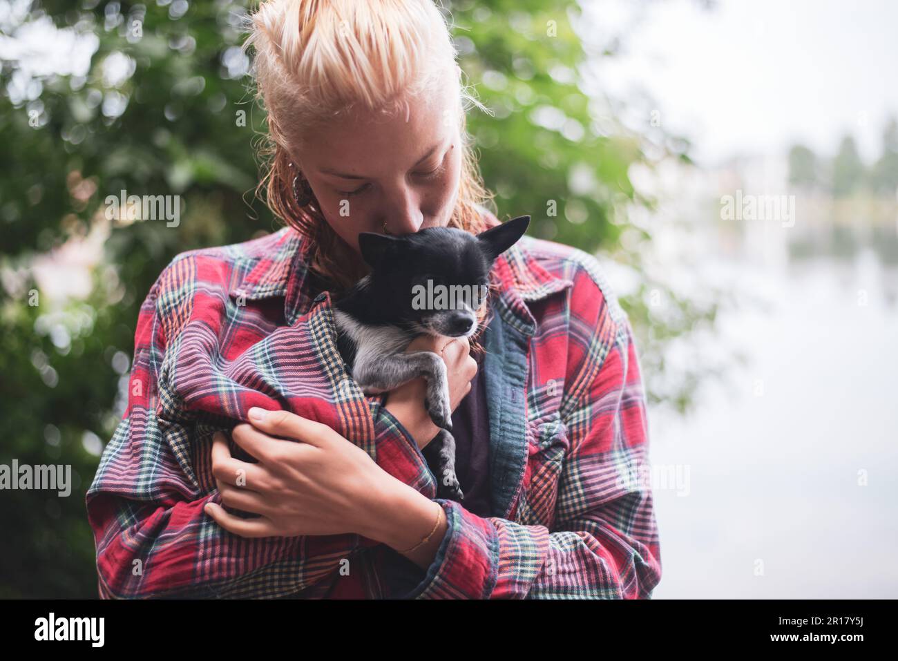 la donna bionda vicino al lago e gli alberi verdi bacia il chihuahua piccolo all'interno della parte superiore Foto Stock