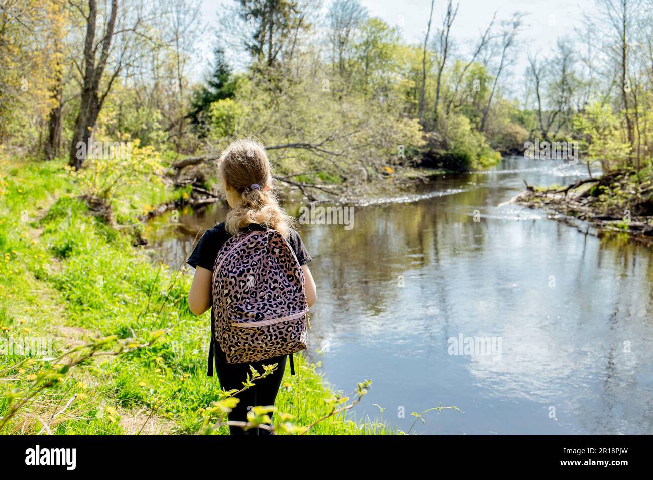 Vista sul retro di una ragazza di 9 anni che cammina da sola nella foresta vicino al fiume con uno zaino all'inizio della primavera. Bellissima natura idilliaca. Foto Stock