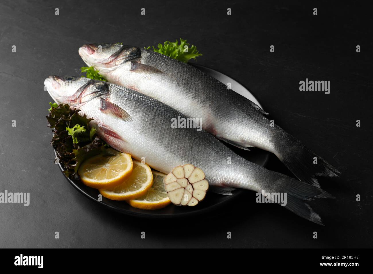 Piatto con pesce fresco crudo di branzino e ingredienti su tavola nera Foto Stock