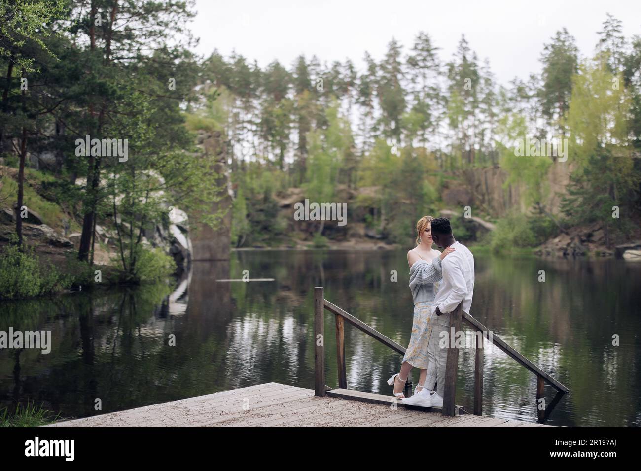 Felice coppia interrazziale si erge su un ponte di legno, si abbraccia e si guarda sullo sfondo del lago e della foresta. Concetto di relazioni d'amore a Foto Stock