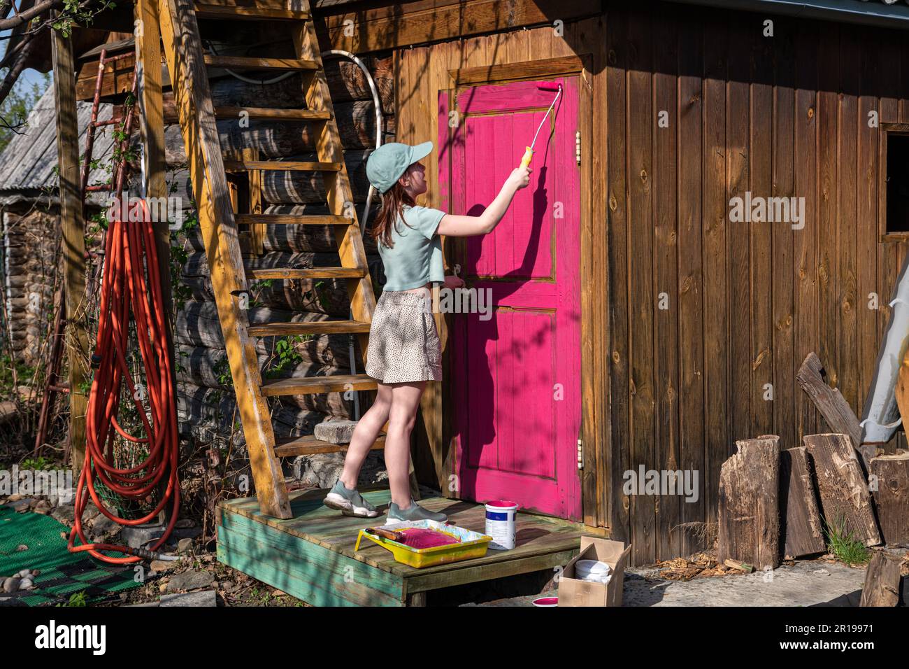 Ragazza carina dipingere la porta di una casa di legno in un chiaro colore viola, rosa, fucsia. casa di campagna, vita di campagna Foto Stock