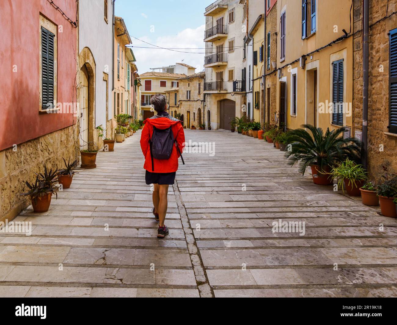 Donna in giacca arancione che cammina per le graziose strade di Alcudia, una città fortificata sulla costa nord-orientale di Maiorca Spagna Foto Stock