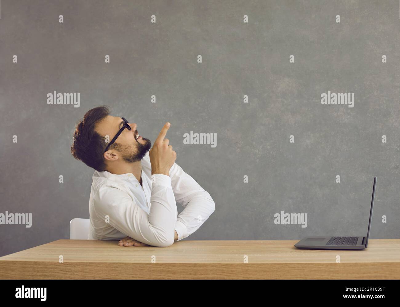 Un giovane uomo con barba e occhiali siede a un tavolo con un computer portatile e mostra il dito indice verso l'alto Foto Stock