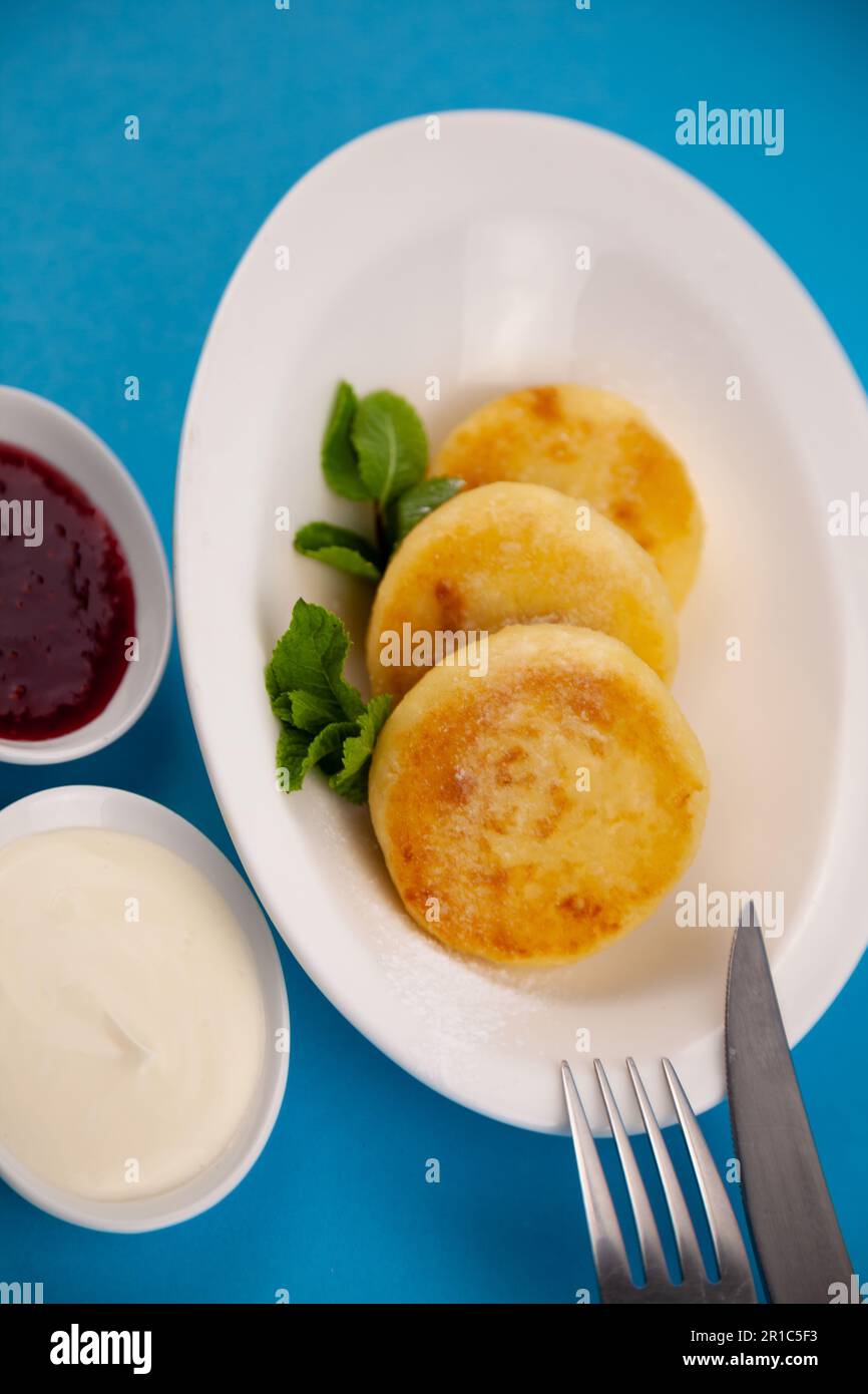 Frittelle di formaggio ricotta rotonda in un piatto bianco su sfondo blu. Cibo prima colazione. Cibo sano. Salsa di panna acida e menta. Primo piano vista dall'alto. Pasto dessert. Frittelle di ricotta al formaggio ricotta. Foto Stock
