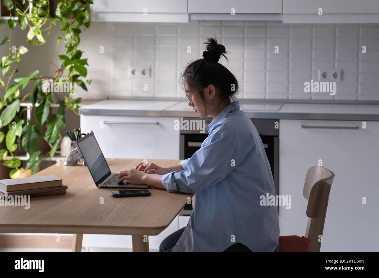 Una donna asiatica focalizzata che lavora da remoto si siede al tavolo con un computer portatile in cucina Foto Stock