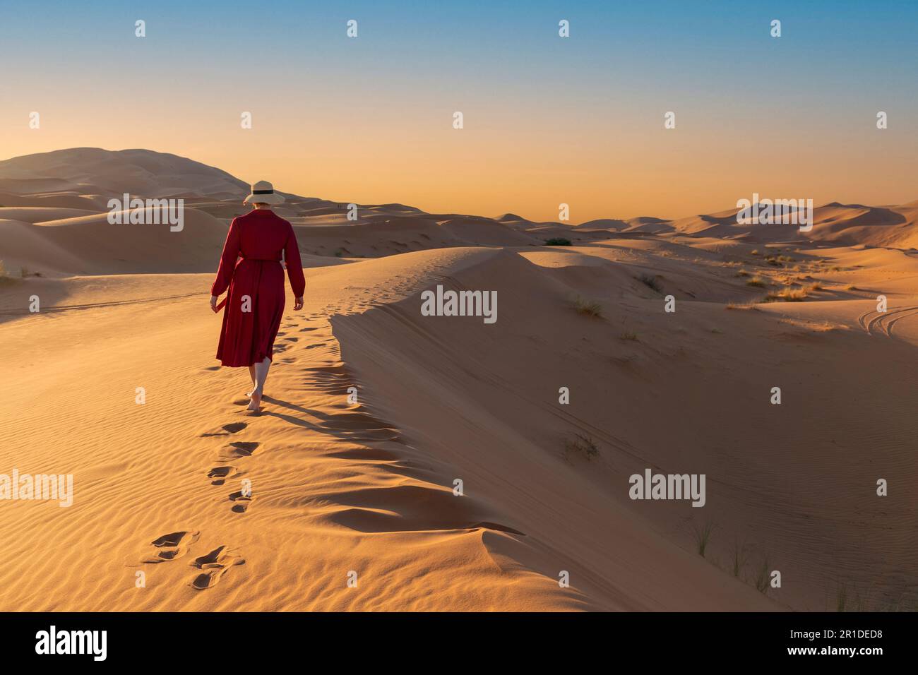 Giovane bella donna in vestito rosso cammina lungo dune di sabbia raggiungendo la cima da sola Foto Stock