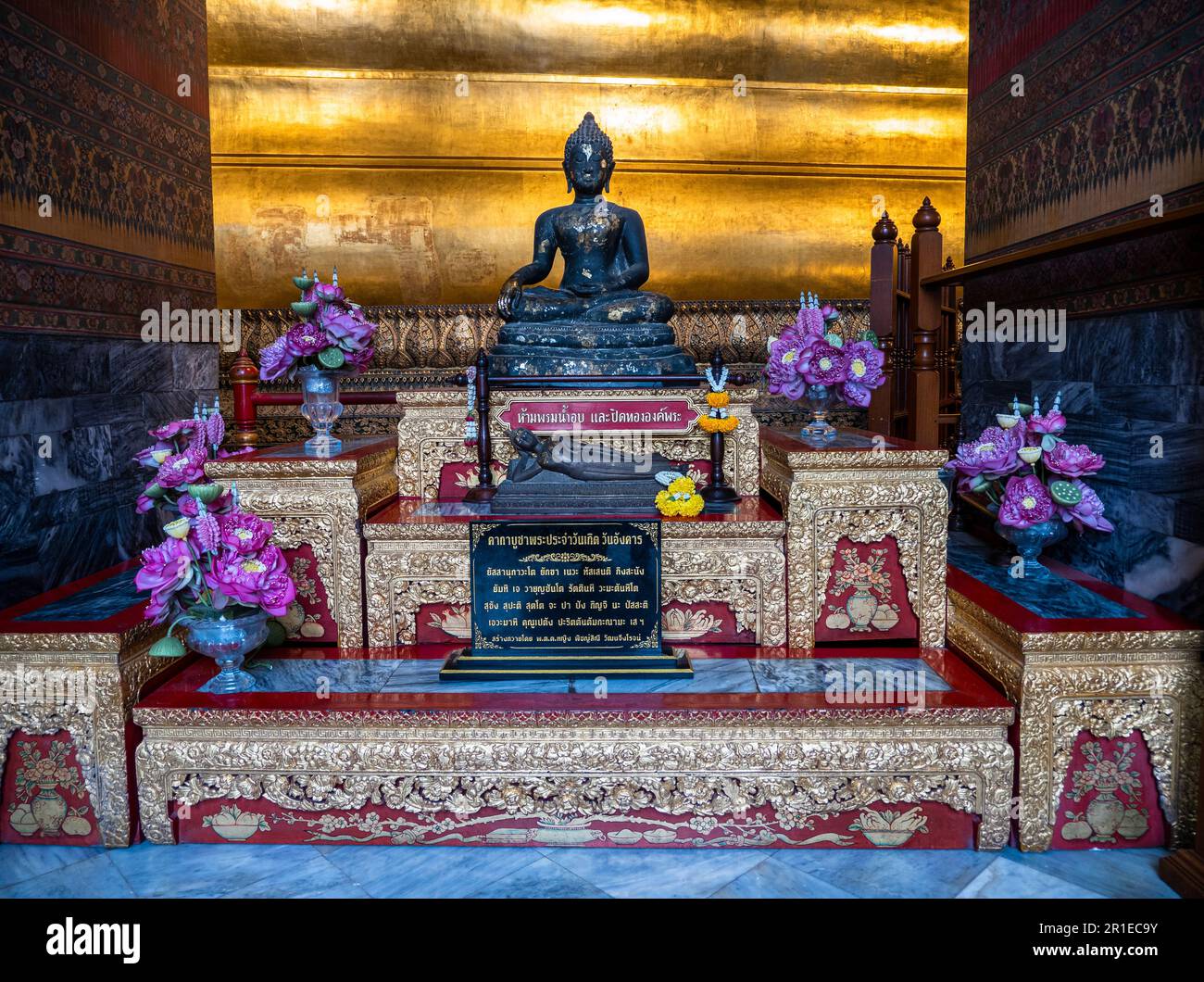 Una statua nera di Buddha con offerte floreali davanti ad una sezione del Buddha sdraiato dorato al tempio di Wat Pho a Bangkok, Thailandia. Foto Stock