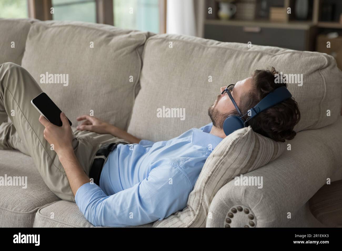 Uomo giovane e rilassato che indossa cuffie wireless e si appoggia sul divano di casa Foto Stock