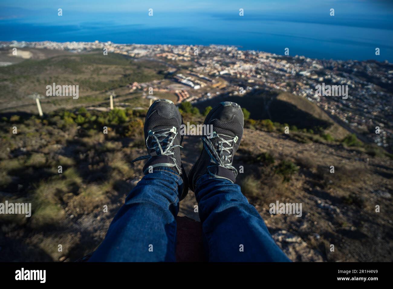 Uomo penzolante le gambe sulla cima del monte Calamorro con una splendida vista sulle montagne e sulla città di Benalmádena, Spagna Foto Stock