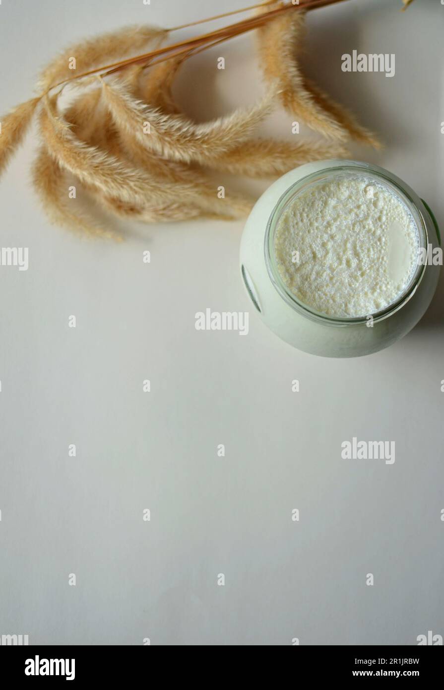 Sul tavolo c'è un vaso di vetro con yogurt bianco fatto in casa. C'è un fiore bianco e spighe di mais sul tavolo. Yogurt greco naturale con schiuma di latte. Foto Stock