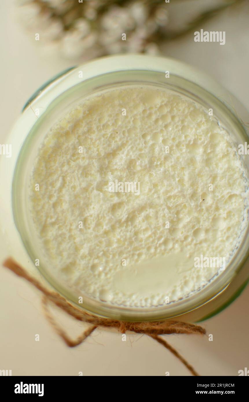 Sul tavolo c'è un vaso di vetro con yogurt bianco fatto in casa. C'è un fiore bianco e spighe di mais sul tavolo. Yogurt greco naturale con schiuma di latte. Foto Stock