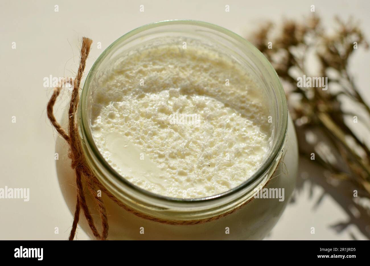 Sul tavolo c'è un vaso di vetro con yogurt bianco fatto in casa. C'è un fiore bianco e spighe di mais sul tavolo. Yogurt greco naturale con schiuma di latte. Foto Stock