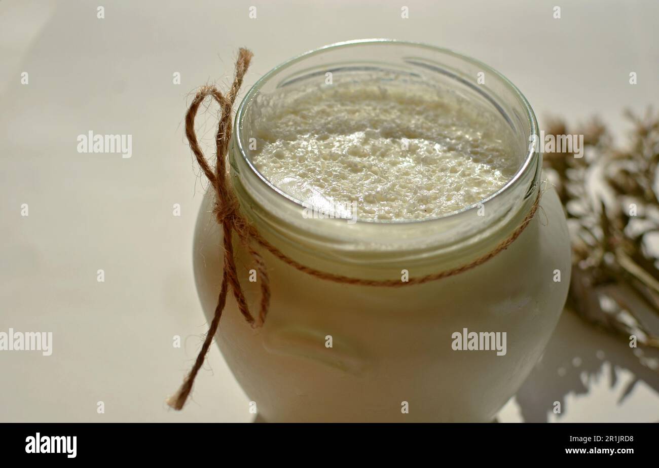 Sul tavolo c'è un vaso di vetro con yogurt bianco fatto in casa. C'è un fiore bianco e spighe di mais sul tavolo. Yogurt greco naturale con schiuma di latte. Foto Stock