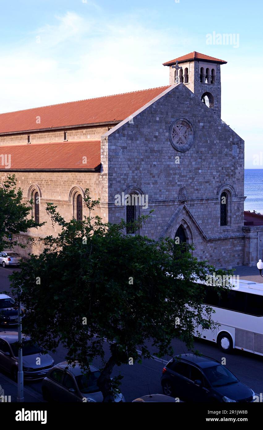 Chiesa cattolica in stile gotico con un campanile, una croce e un tetto rosso. Alberi sulla strada e Cars.Temple sul mare, Piazza Mandraki Foto Stock