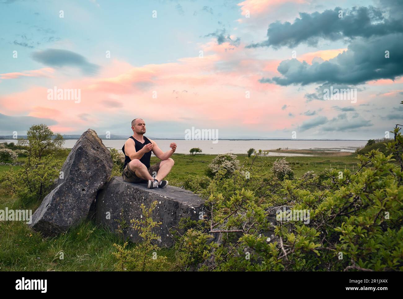 Splendido scenario naturalistico con uomo in camicia senza maniche e pantaloncini camo meditati sulla roccia sul lago Corrib a Galway, Irlanda Foto Stock