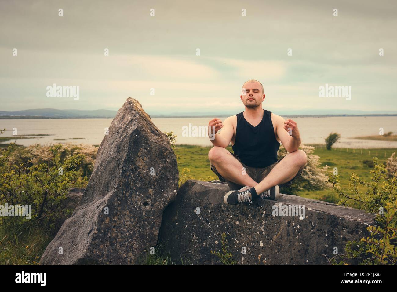 Splendido scenario naturalistico con uomo in camicia senza maniche e pantaloncini camo meditati sulla roccia sul lago Corrib a Galway, Irlanda Foto Stock