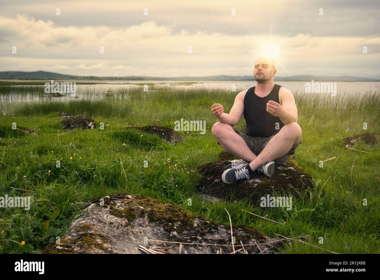 Splendido scenario naturalistico con uomo in camicia senza maniche e pantaloncini camo meditati sulla roccia sul lago Corrib a Galway, Irlanda Foto Stock
