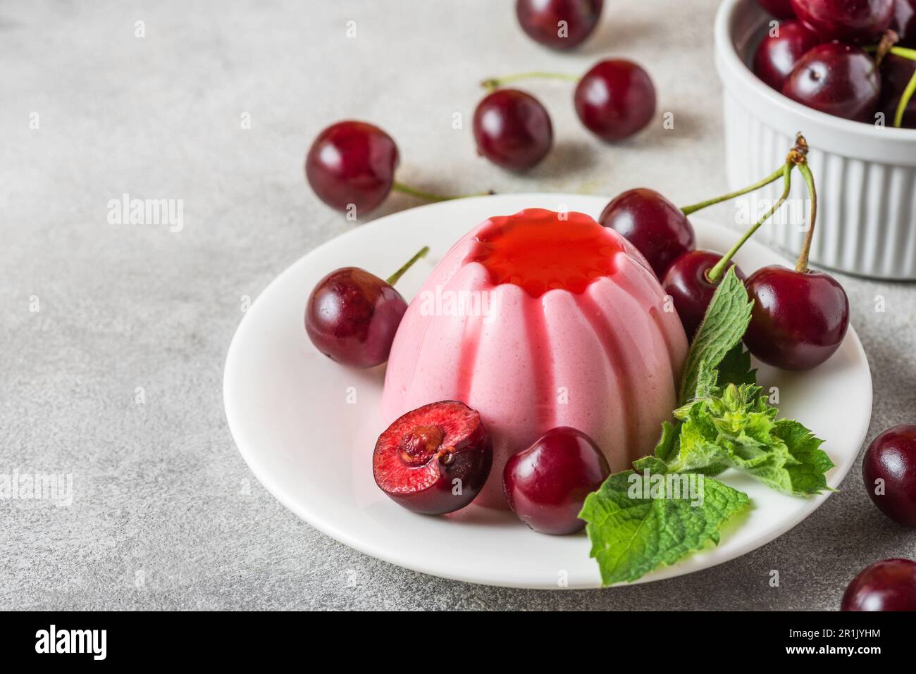 Delizioso dessert di panna cotta alle ciliegie con frutti di bosco freschi, salsa di frutta e menta in un piatto su un tavolo grigio Foto Stock