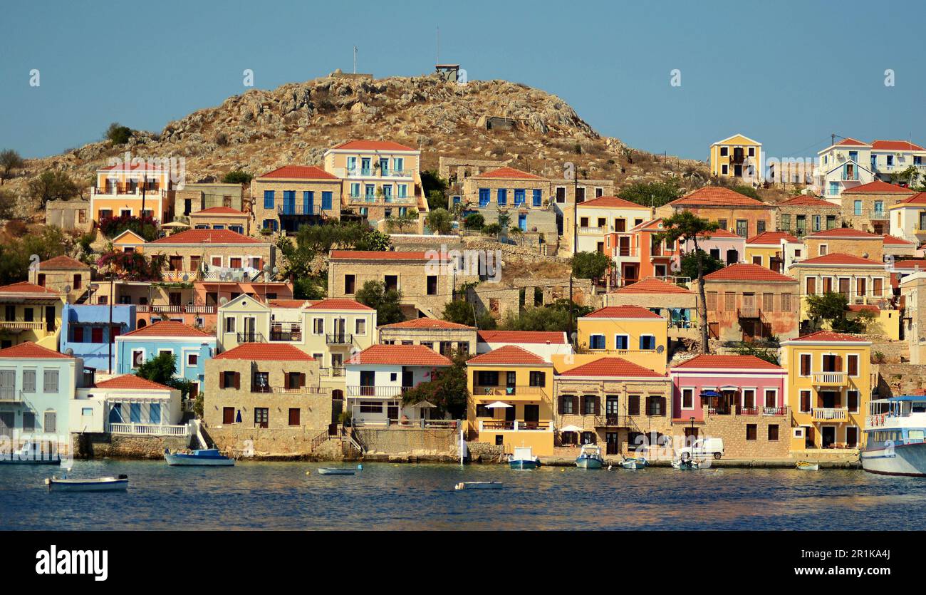 Case colorate sulle pendici di una collina nel porto dell'isola di Halki in Grecia. Piccole barche da pesca sono ormeggiate al largo della costa, nelle acque turchesi. Foto Stock