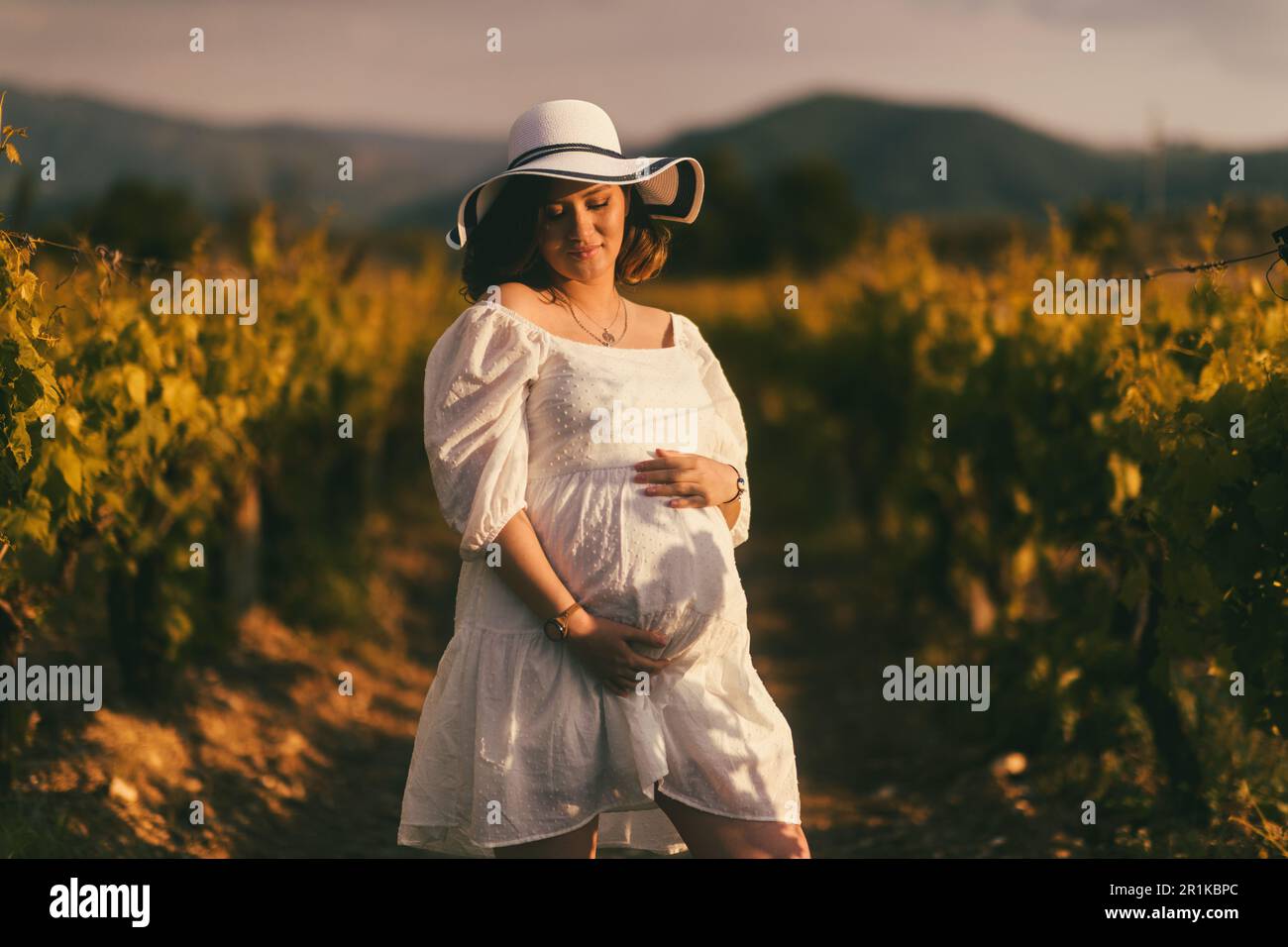 Un ritratto di una donna incinta in abito bianco e cappello, tenendo dolcemente il ventre, nella cornice di una vigna, irradiando maternità e attesa Foto Stock