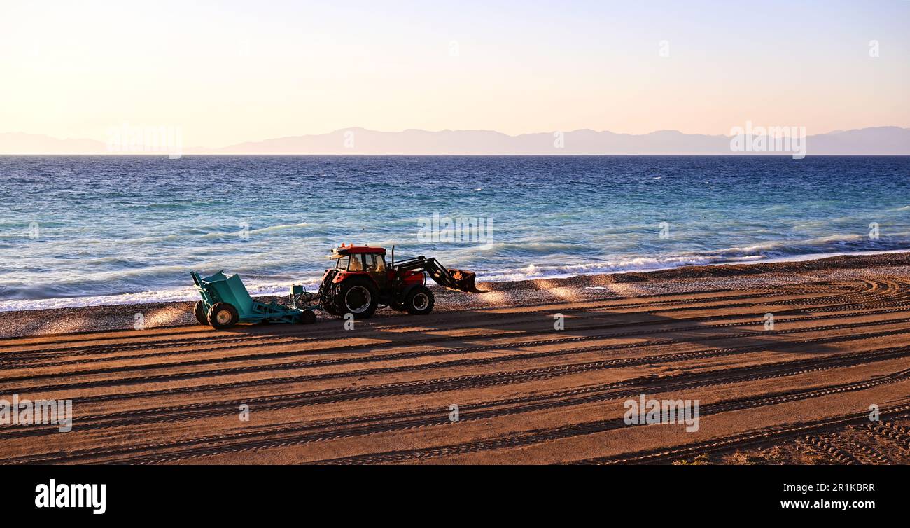 Trattore con rimorchio per la pulizia della spiaggia dai detriti invernali. Il trattore lavora in riva al mare, vicino alle onde. Il trattore depone sabbia sulla spiaggia. Foto Stock