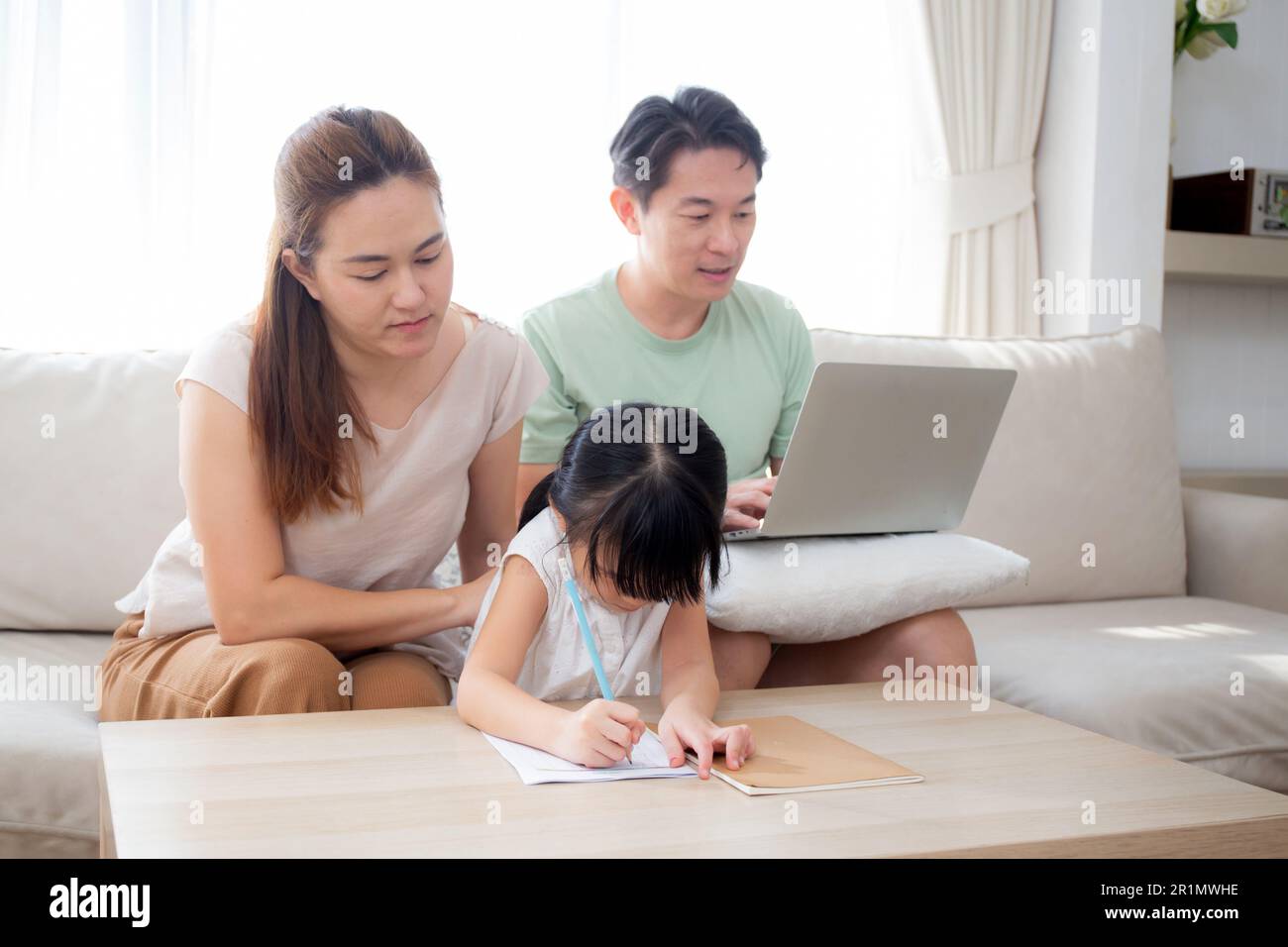 Famiglia felice con la madre che insegna i compiti a casa con la figlia e il padre che lavora con il portatile sul divano nel soggiorno di casa, mamma che spiega il lavoro scolastico con Foto Stock