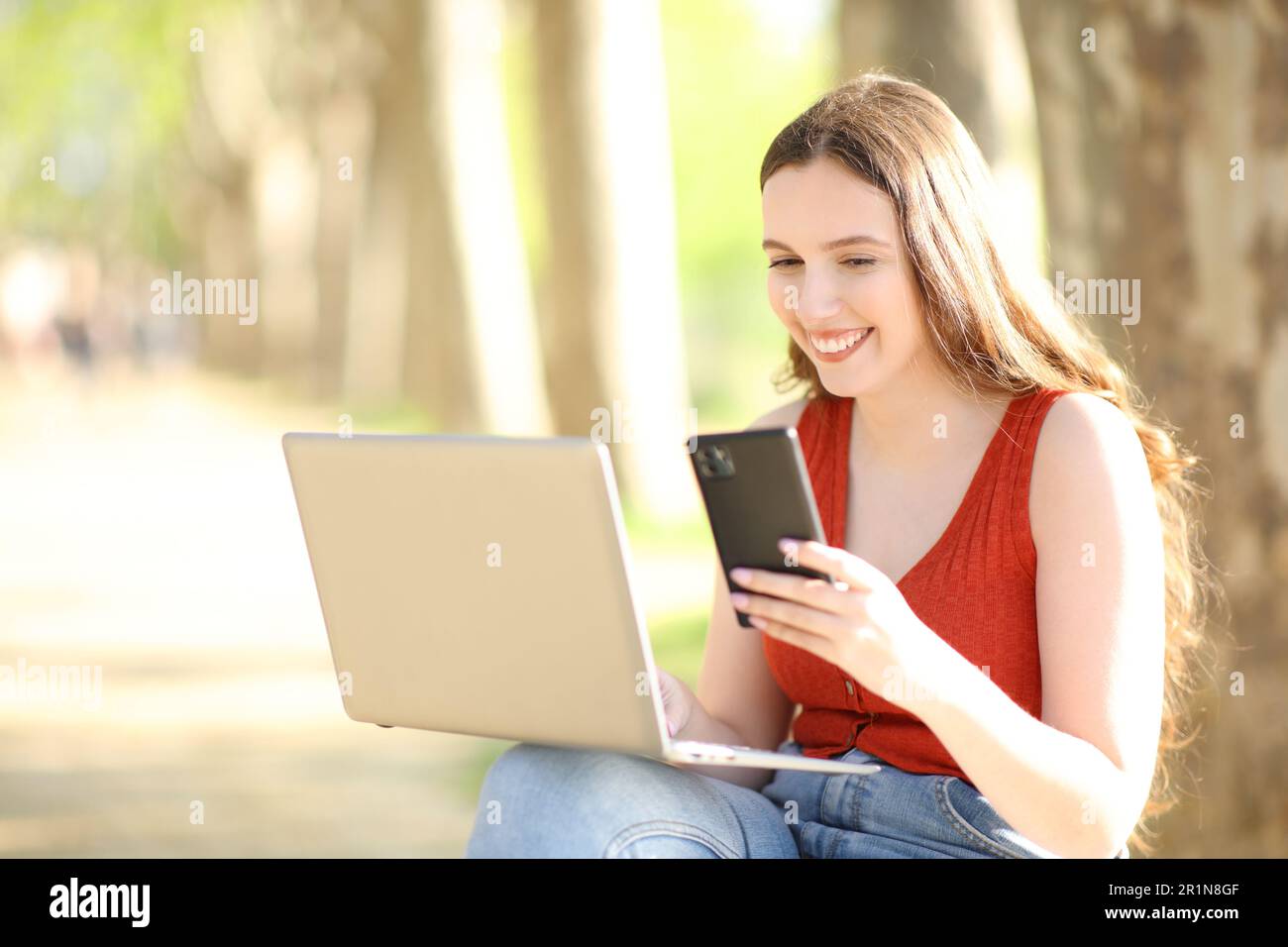 Donna felice che usa il portatile e il telefono cellulare seduto in un parco in estate Foto Stock