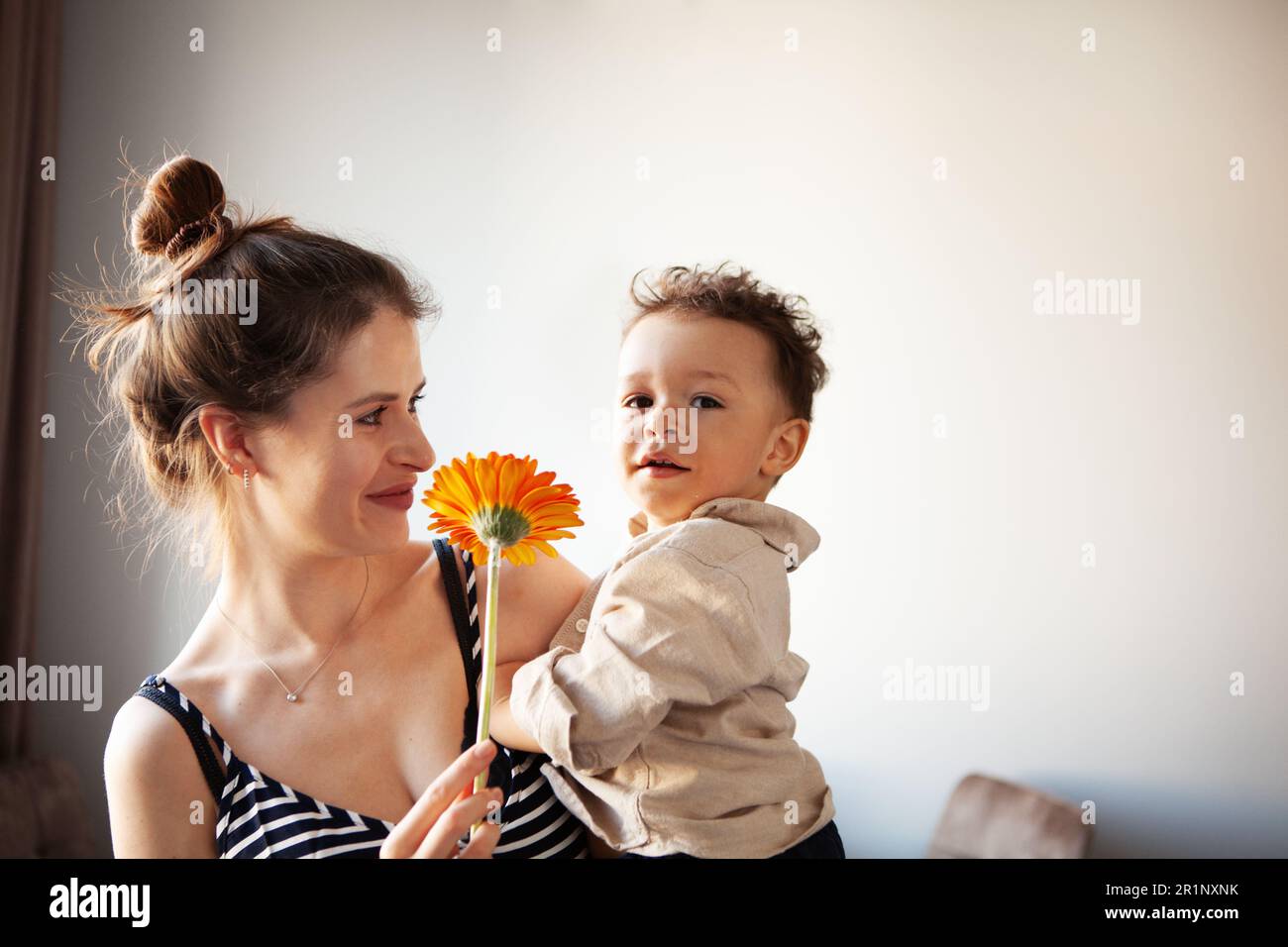 Mamma che tiene in mano il bambino, che guarda nella fotocamera mentre si aggrappano i fiori. Vita reale e autenticità su sfondo chiaro. Foto Stock