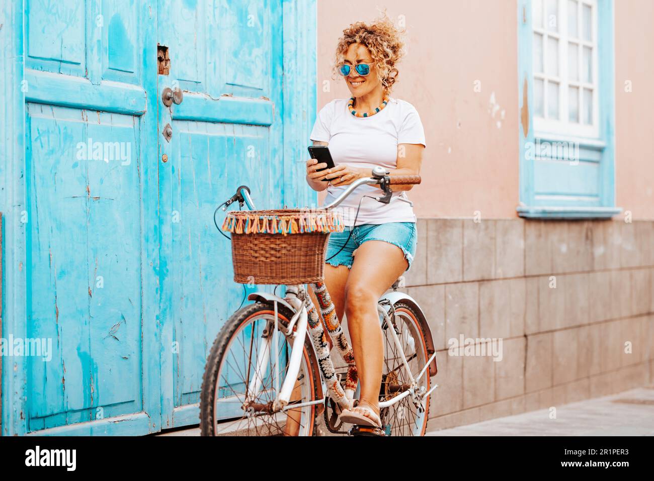 Una giovane donna matura in attività di svago all'aperto in bicicletta e utilizzando il telefono cellulare fuori casa in strada. Primavera attiva verde trasporto bicicletta persone femmina. Donna felice utilizzando il cellulare Foto Stock