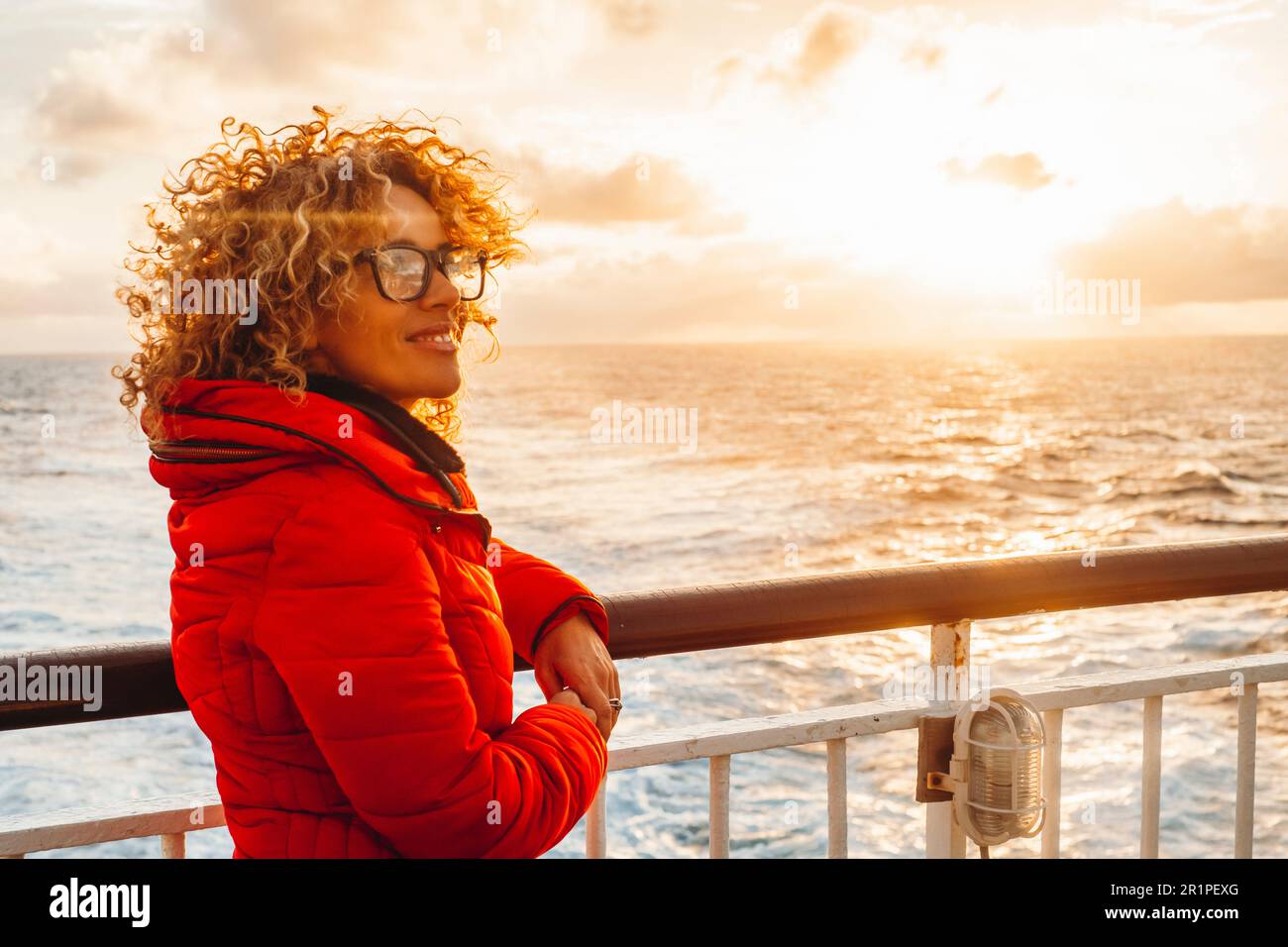 Ritratto di una giovane donna allegra adulta godendo il tramonto fuori viaggiare l'oceano su un traghetto da solo nave da crociera. Stile di vita dei viaggiatori, giacca rossa. Sfondo arancione cielo caldo. Trasporto Foto Stock