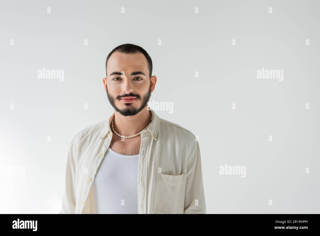 Ritratto di giovane uomo omosessuale a pelo corto e bearato in collana di perle e abiti casual guardando la macchina fotografica mentre si è in piedi isolato sul grigio Foto Stock