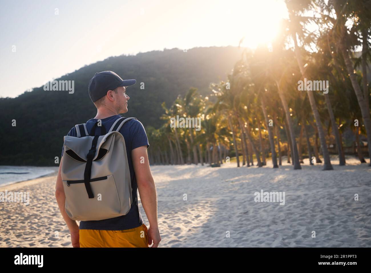 Vista posteriore del turista con zaino a piedi sulla bellissima spiaggia di sabbia con palme e guardare il tramonto. Foto Stock