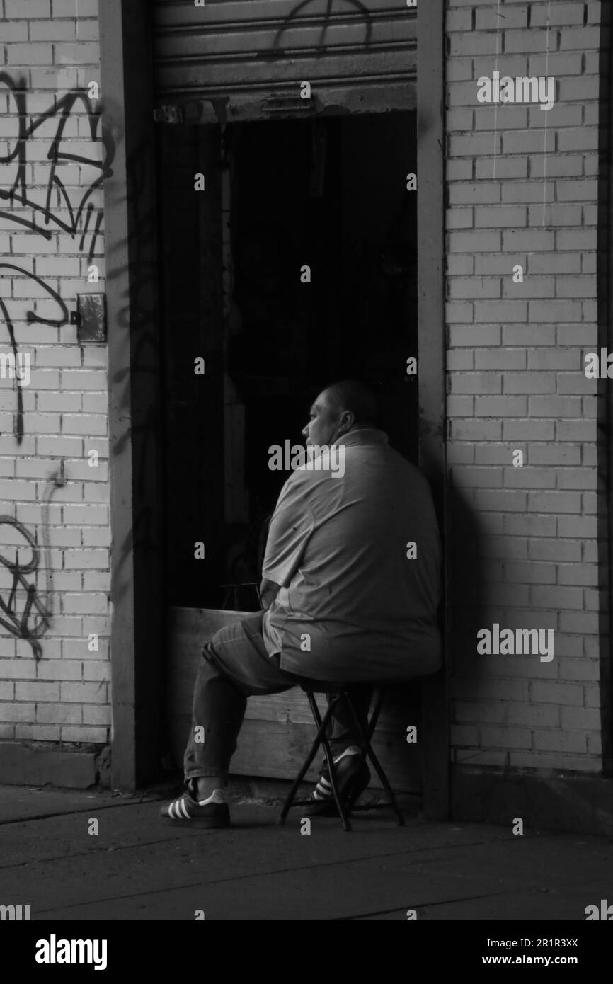Un'immagine in scala di grigi di un uomo seduto su una sedia di fronte a una parete con un vivace murale di graffiti. Foto Stock