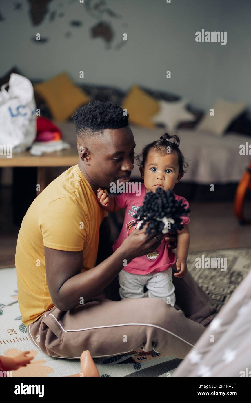 Il giovane uomo africano gioca con la sua figlia del bambino dal matrimonio interracial nella stanza fra i giocattoli sul pavimento. Concetto di famiglia interracial. Foto Stock