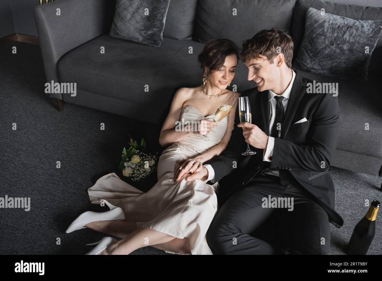 sposi felici in eleganti bicchieri di champagne da fiancare in abito celebrando il loro matrimonio vicino al bouquet nuziale e alla bottiglia sul pavimento dopo il matrimonio Foto Stock