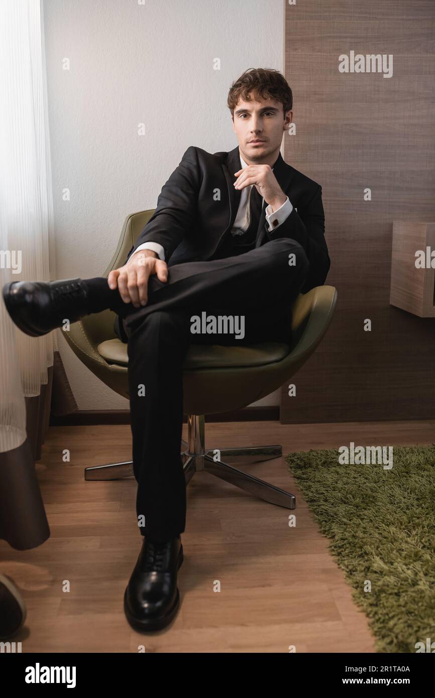 giovane uomo in abito nero formale con cravatta e scarpe classiche seduto su una comoda poltrona e guardando la macchina fotografica il giorno del matrimonio, posa in un hotel moderno Foto Stock