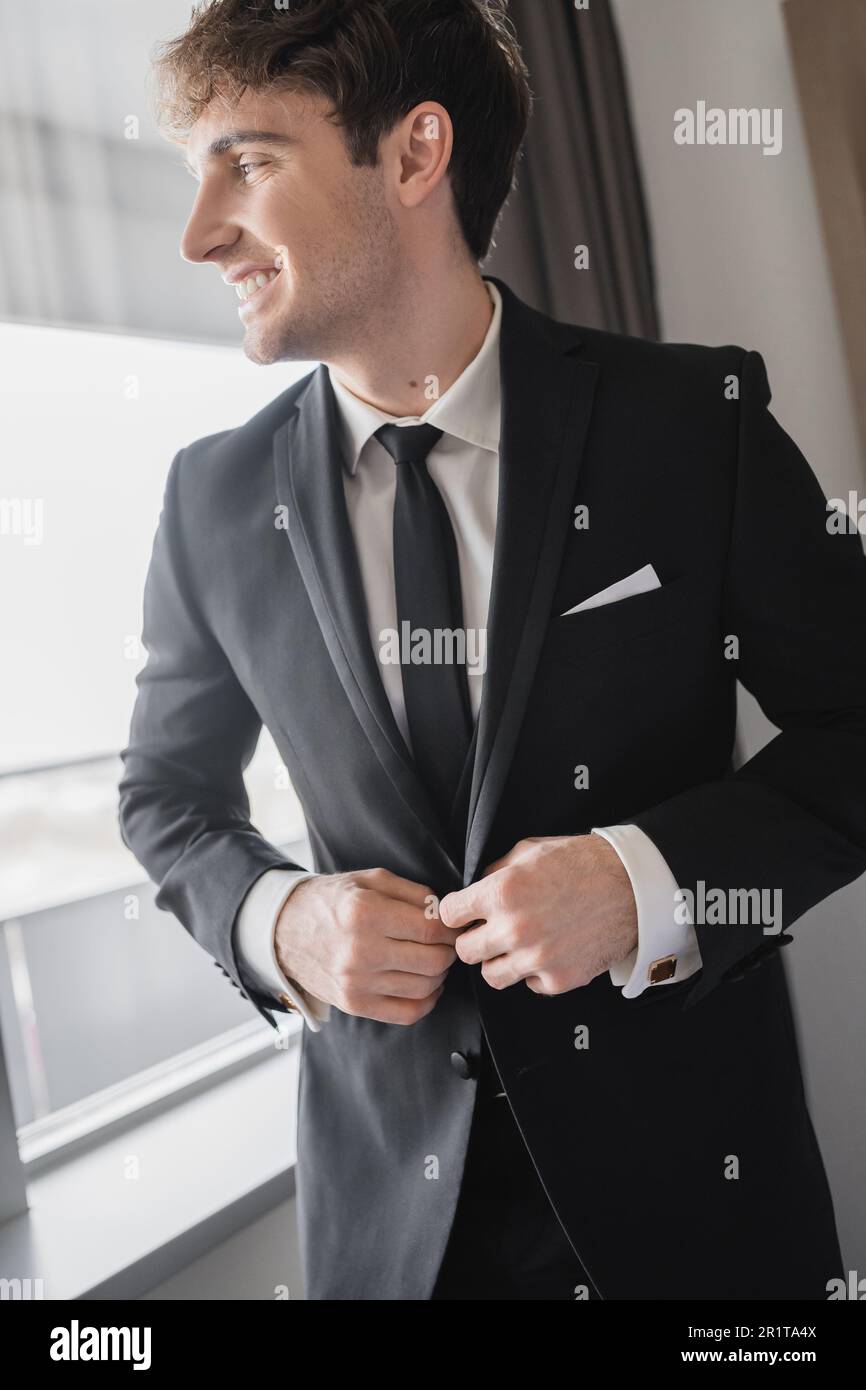 uomo felice in classico abbigliamento formale con cravatta nera e camicia bianca blazer bottoning e in piedi nella moderna camera d'albergo vicino alla finestra, sposo il giorno del matrimonio, Foto Stock