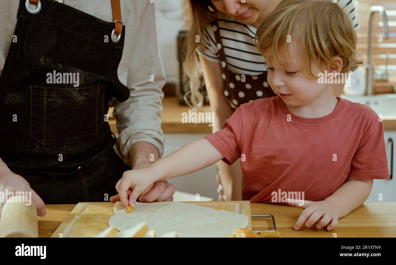 Felice giovane famiglia godendo preparare pasta di torta o dolci in cucina moderna insieme, genitori molto felici che insegnano a piccolo figlio come cucinare panetteria a casa Foto Stock