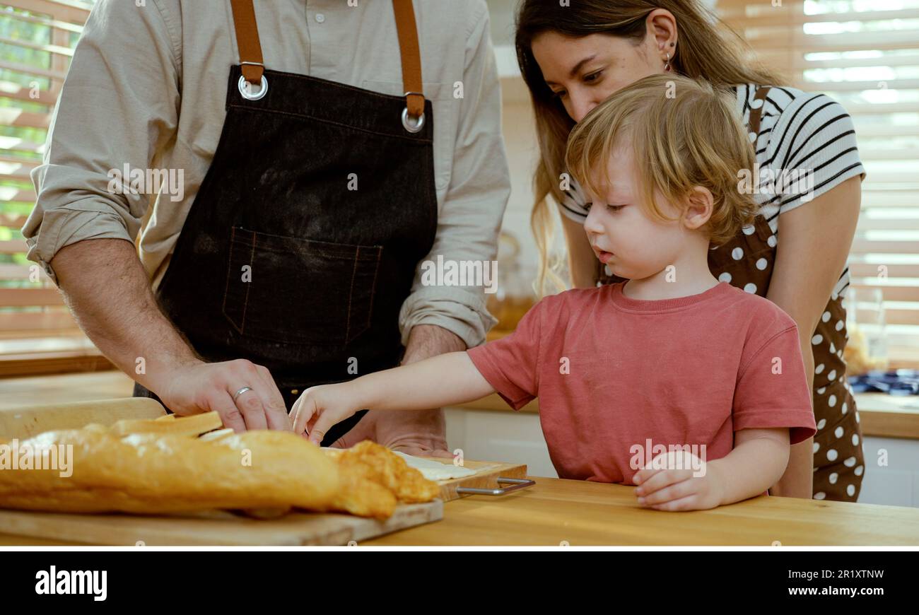 Felice giovane famiglia godendo preparare pasta di torta o dolci in cucina moderna insieme, genitori molto felici che insegnano a piccolo figlio come cucinare panetteria a casa Foto Stock