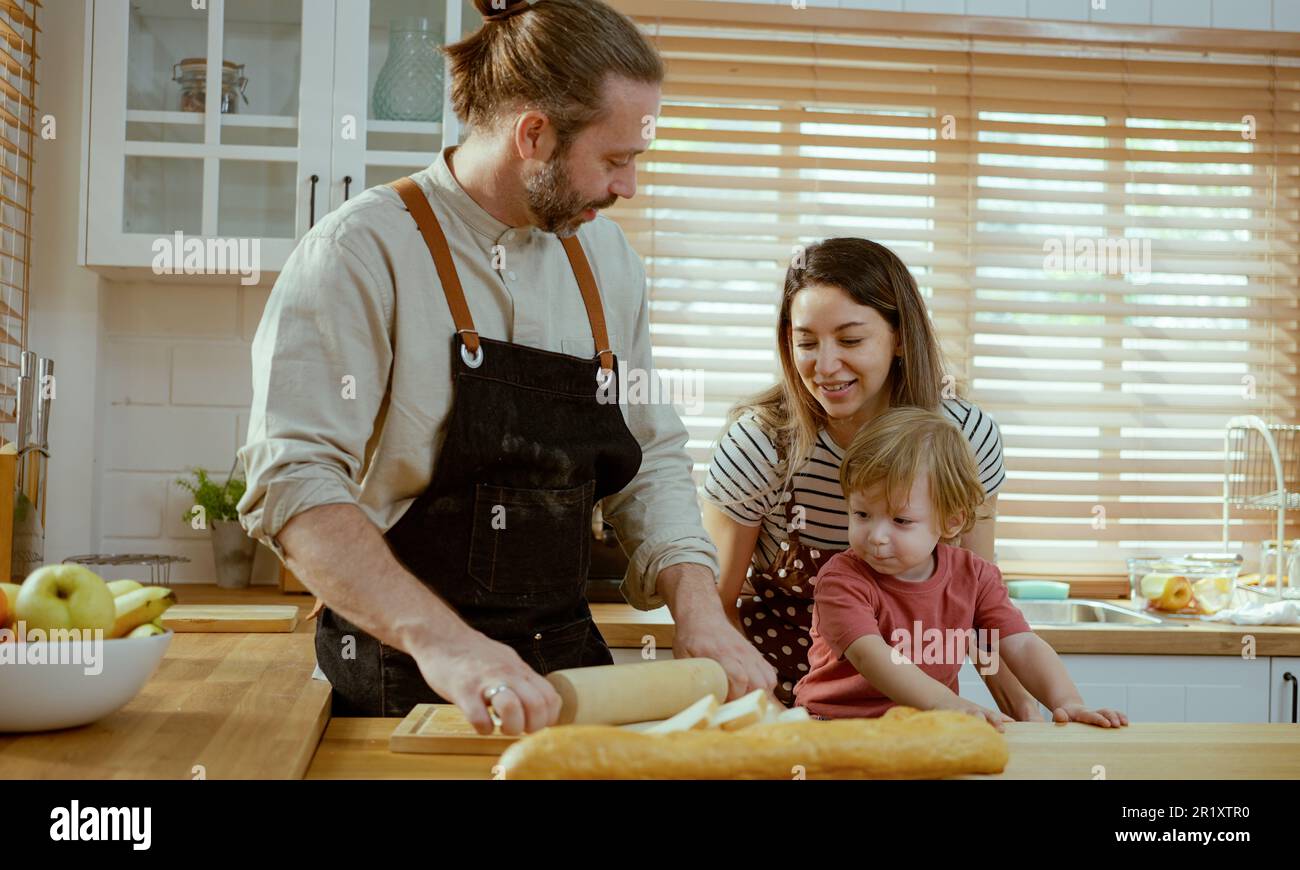 Felice giovane famiglia godendo preparare pasta di torta o dolci in cucina moderna insieme, genitori molto felici che insegnano a piccolo figlio come cucinare panetteria a casa Foto Stock