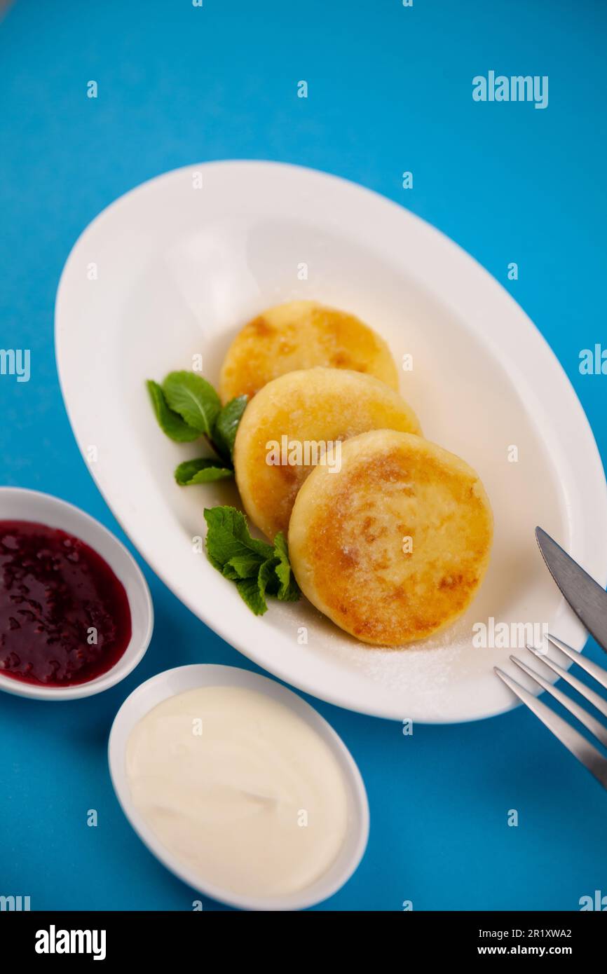 Frittelle di formaggio ricotta rotonda in un piatto bianco su sfondo blu. Cibo prima colazione. Cibo sano. Salsa di panna acida e menta. Primo piano vista dall'alto. Pasto dessert. Frittelle di ricotta al formaggio ricotta. Foto Stock