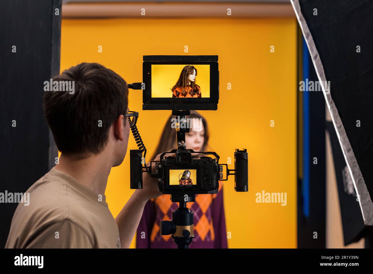 Vista di un fotografo professionista che scatta una giovane donna utilizzando una fotocamera con display esterno, sfondo giallo Foto Stock