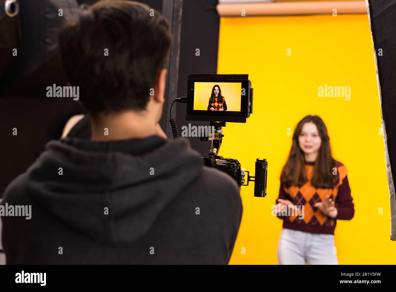 Vista di un fotografo professionista che scatta una giovane donna utilizzando una fotocamera con display esterno, sfondo giallo Foto Stock