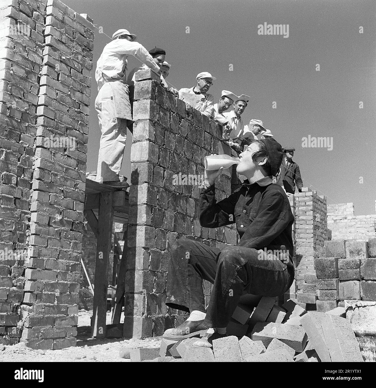 Negli anni '1950s. Una giovane apprendista di mattoni beve da una bottiglia di latte al sole durante una pausa al lavoro. Il team di lavoratori è visto sul cantiere dietro di lei. A questo punto il latte è stato venduto e consegnato in bottiglie di vetro o si potrebbe comprare il latte per portare a casa dal negozio nel proprio biberon. Svezia 1951. Kristoffersson rif BB94-9 Foto Stock