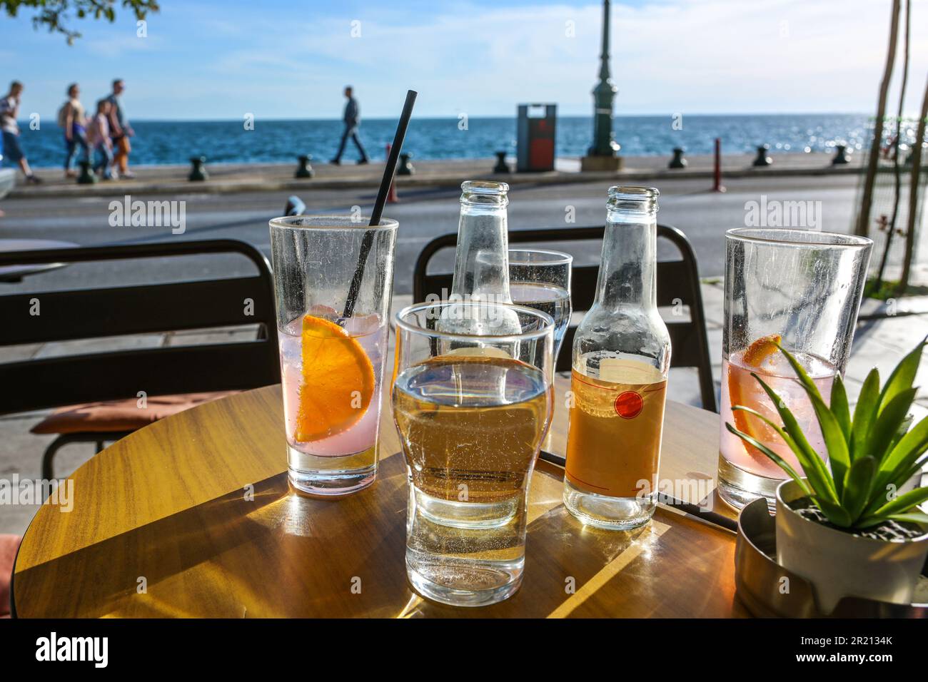 Bevande rinfrescanti in bicchieri da bere e bottiglie su un tavolo in un caffè di strada sul mare, persone blurry a piedi sul lungomare su un s. Foto Stock