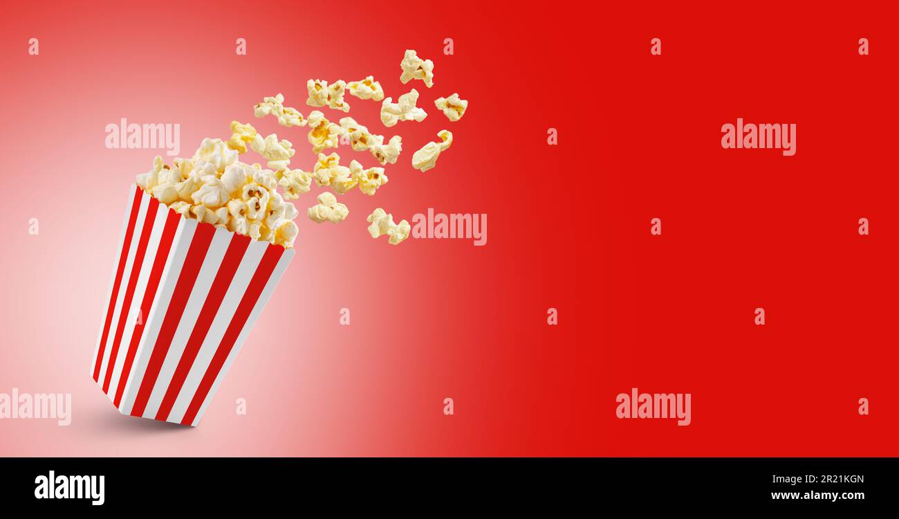 Popcorn che vola fuori dalla scatola di carta a strisce bianche rosse isolato su sfondo rosso con spazio di copia. Spruzzi, levitazione di grani di popcorn. Foto Stock