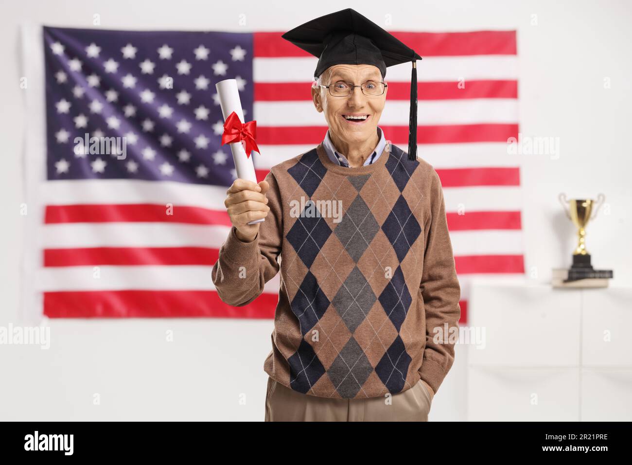 Uomo anziano eccitato con un cappello di laurea e un diploma in posa davanti ad una bandiera degli Stati Uniti Foto Stock
