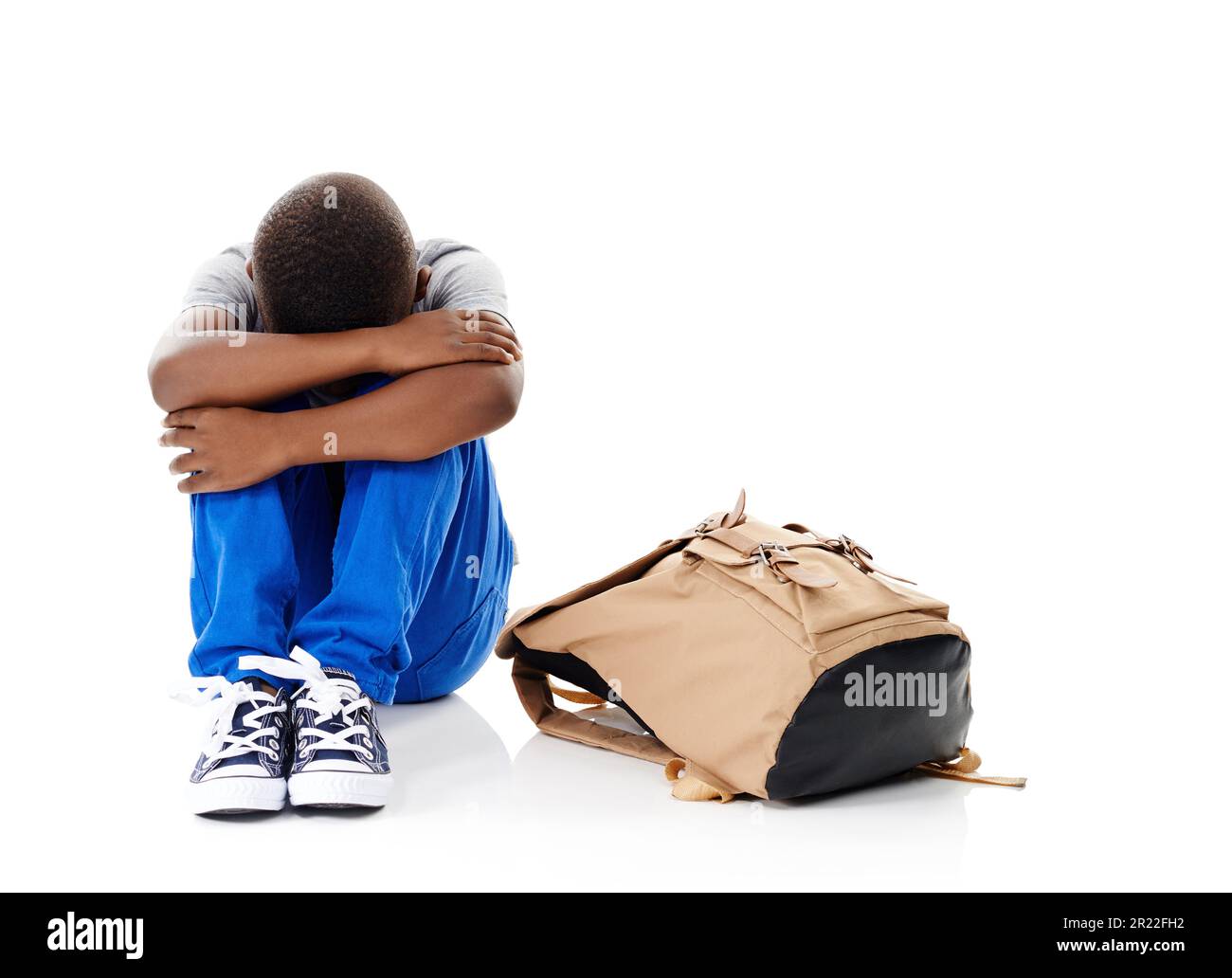 Non voglio. Studio shot di un ragazzino con la testa sepolta in ginocchio seduto accanto alla sua borsa contro uno sfondo bianco. Foto Stock