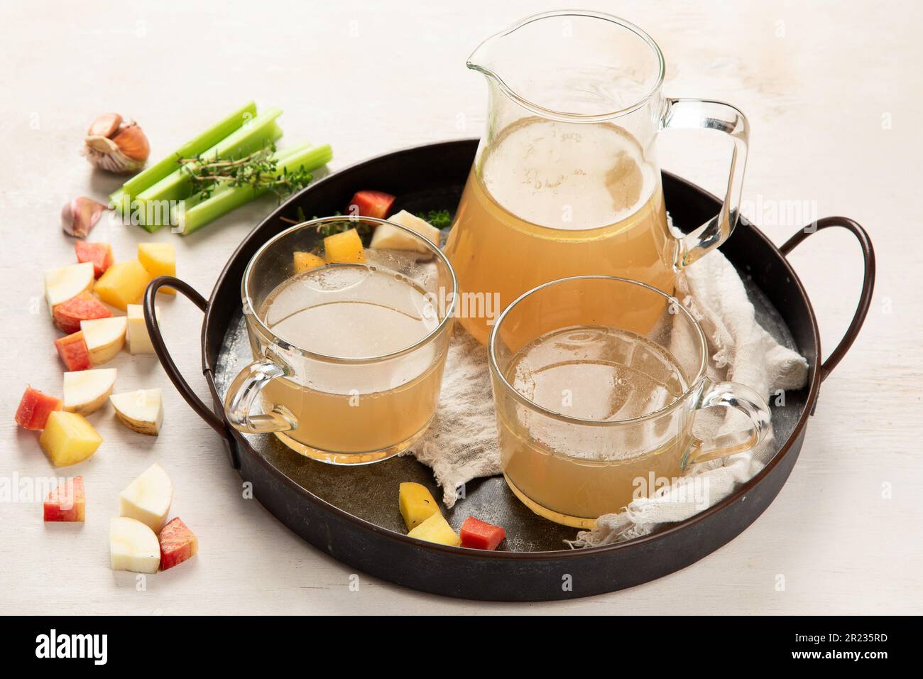 Due tazze e tazza di vetro di brodo di ossa di pollo fatto in casa su sfondo bianco. Brodo osseo, brodo osseo, spicchi d'aglio ed erbe aromatiche. Foto Stock