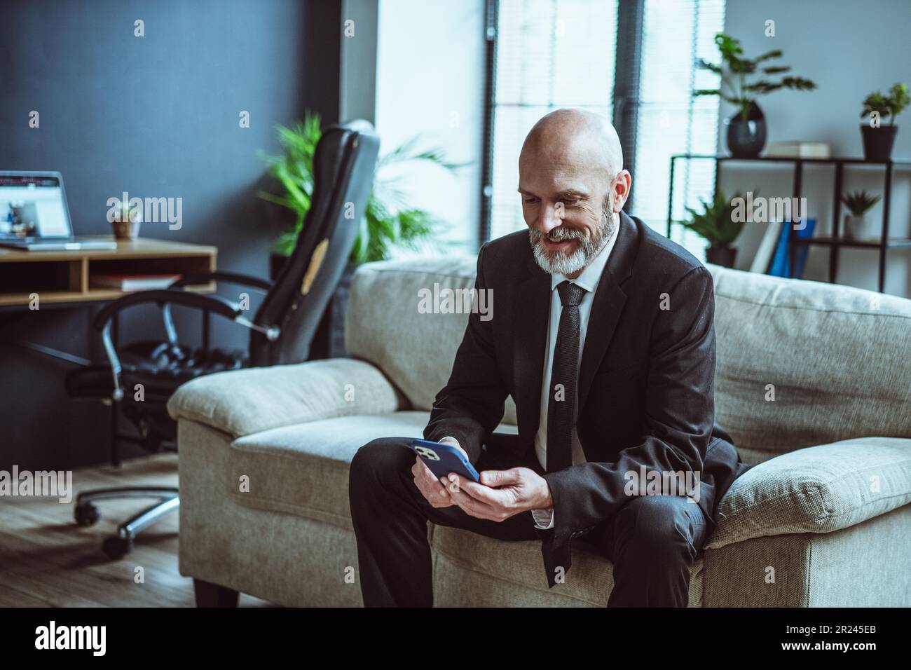 Uomo d'affari anziano vestito di nero, con un sorriso caldo e sportivo, mentre navighi su internet al telefono. Uomo maturo, con la barba d'argento distinta, visto che riposa sul divano in loft ufficio. . Foto di alta qualità Foto Stock